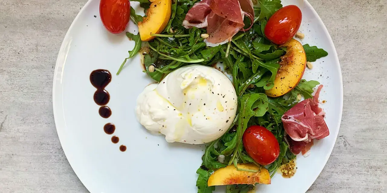 Burrata mit Rucola, Tomaten, Pfirsichspalten und Prosciutto auf einem Teller.