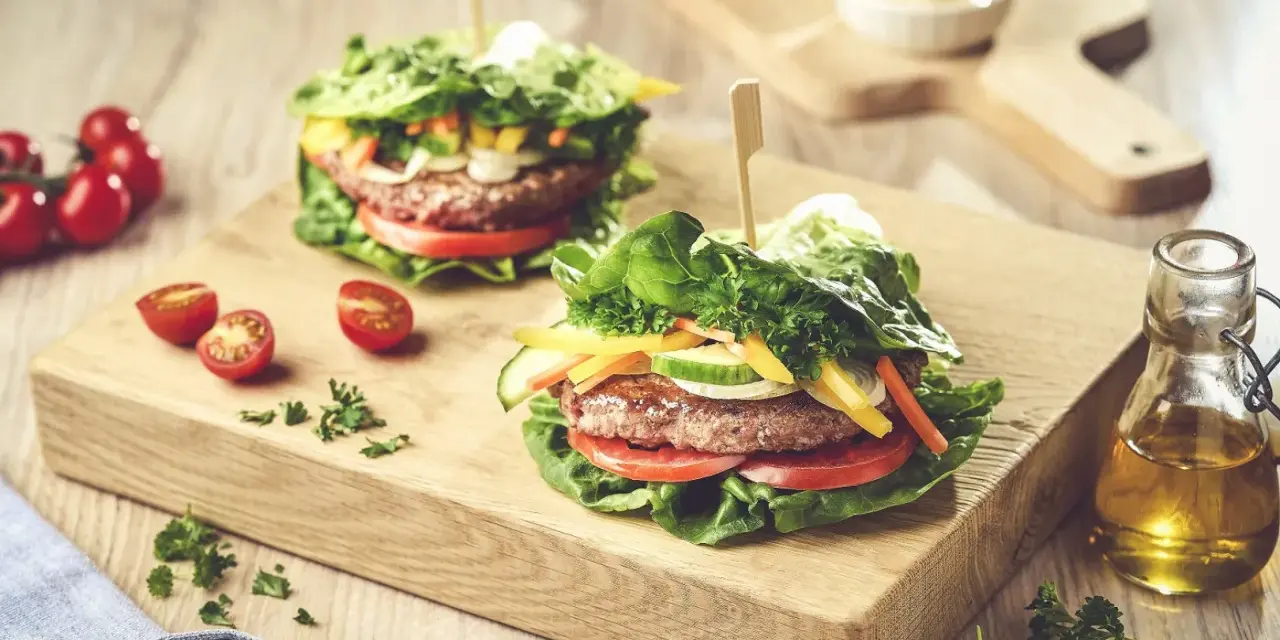 Dwa soczyste burgery w sałacie, z pomidorami, ogórkiem i papryką, na drewnianej desce.