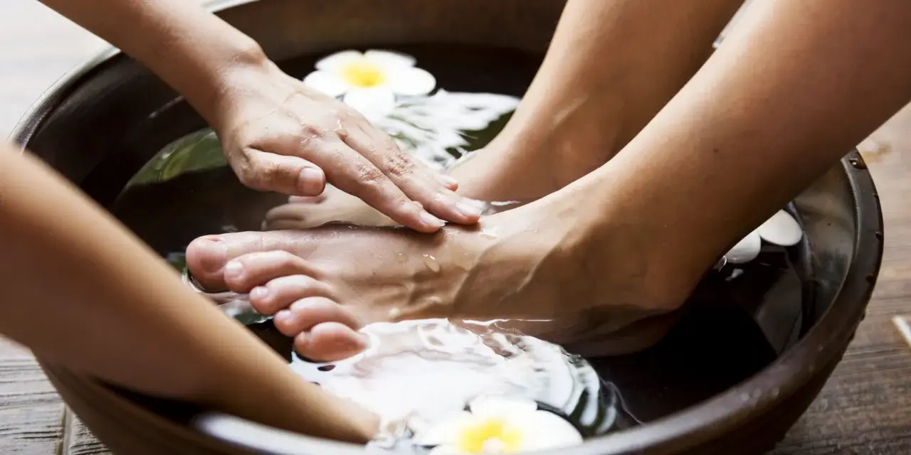 Soaking feet in a warm bath with Epsom salt for feet infection. A hand gently massages the foot, surrounded by water and white flowers.