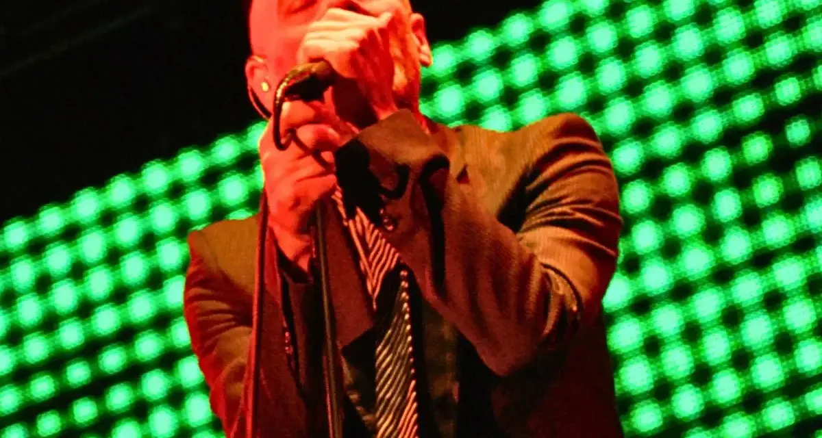 Wokalista REM, Michael Stipe, śpiewa na scenie, gdy światła migoczą na zielono.