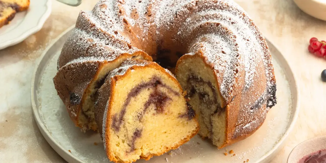 Ein Gugelhupf mit Marmelade, bestäubt mit Puderzucker. Ein Stück ist bereits angeschnitten und zeigt die köstliche Füllung.