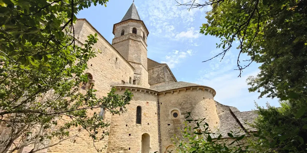 Vue d'une église romane à Villeneuve de Rouergue, encadrée par des arbres verdoyants sous un ciel bleu parsemé de nuages.