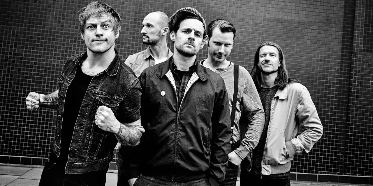 Fünf Männer stehen vor einer gemusterten Wand. Sie sehen aus wie eine energiegeladene Punkband, bereit, die Bühne zu rocken.