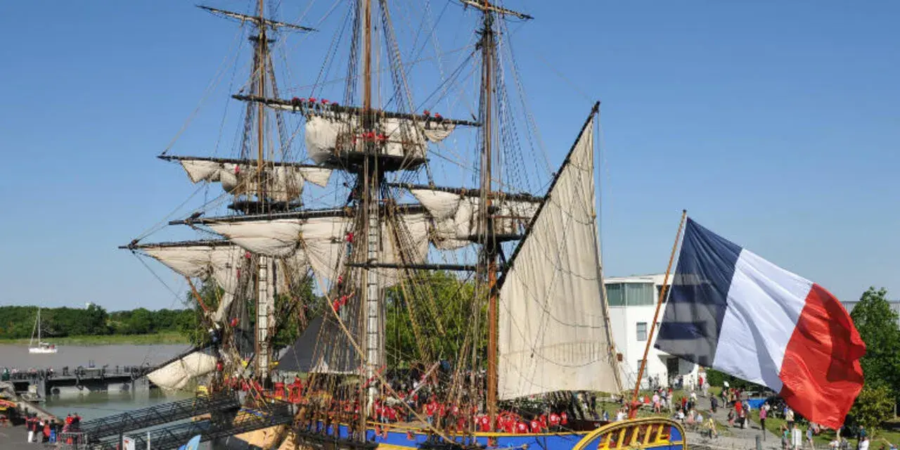 Le majestueux bateau l'Hermione, réplique d'une frégate du XVIIIe siècle, est amarré, pavoisé du drapeau français. Une foule s'est rassemblée pour admirer ce navire historique.