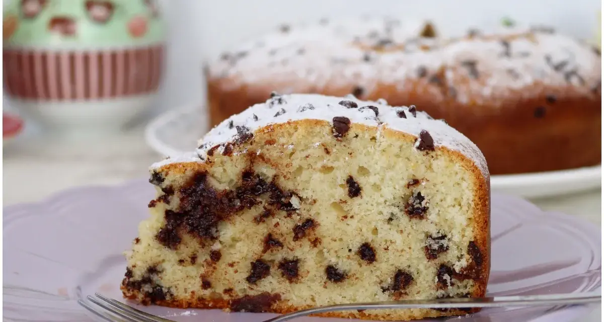 Una fetta di soffice torta con gocce di cioccolato, cosparsa di zucchero a velo, su un piatto lilla con una forchetta.