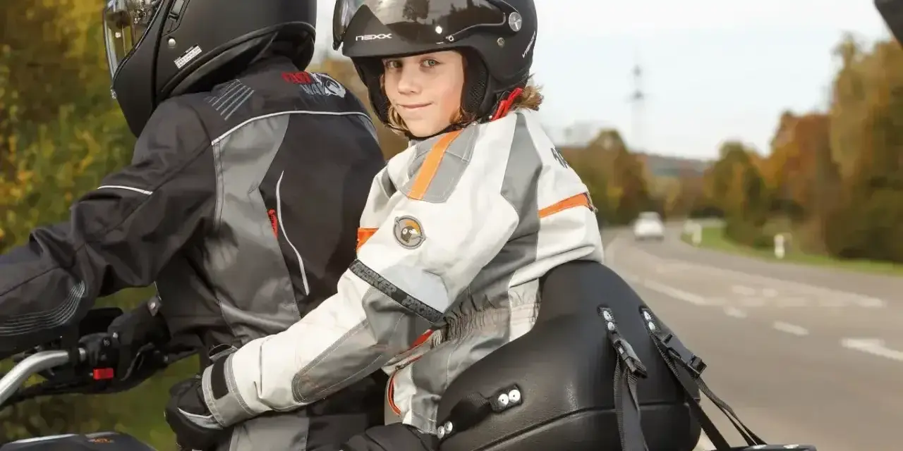 Uśmiechnięte dziecko na motocyklu, siedzące przed dorosłym, w kasku i kurtce motocyklowej.