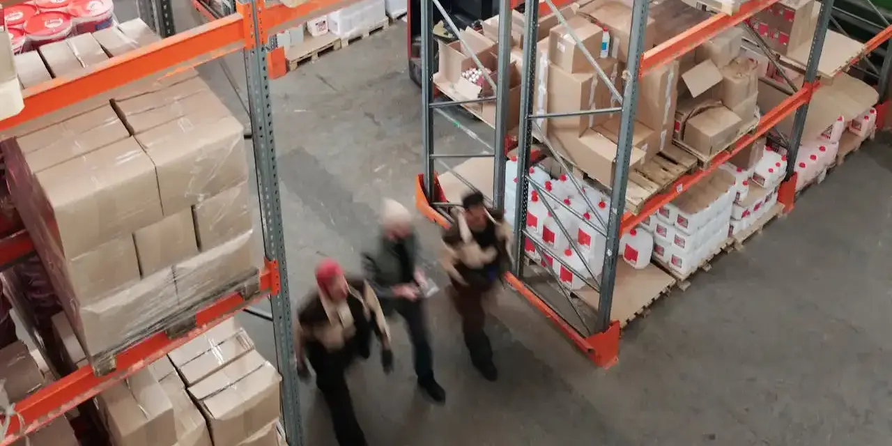 Magazzino con scaffalature piene di scatoloni e taniche. Tre persone camminano velocemente, forse per svolgere mansioni di aiuto magazziniere.