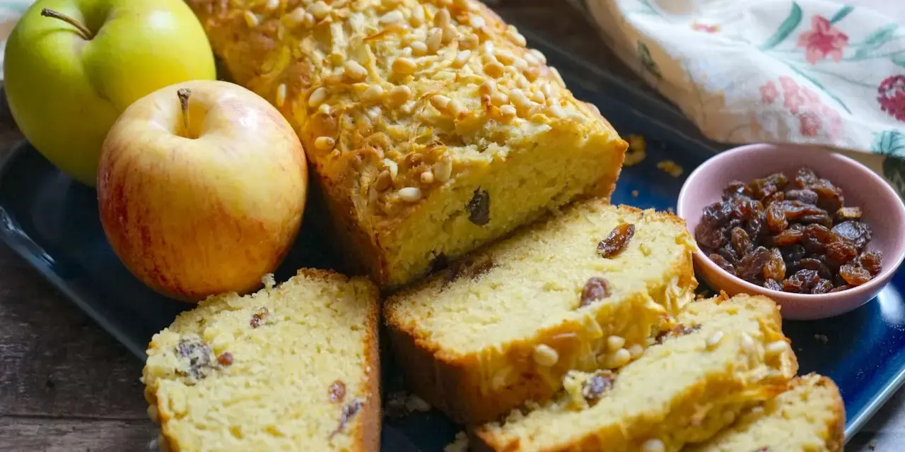 Plumcake alle mele dorato, con pinoli e uvetta, accompagnato da mele fresche e una ciotolina di uvetta.