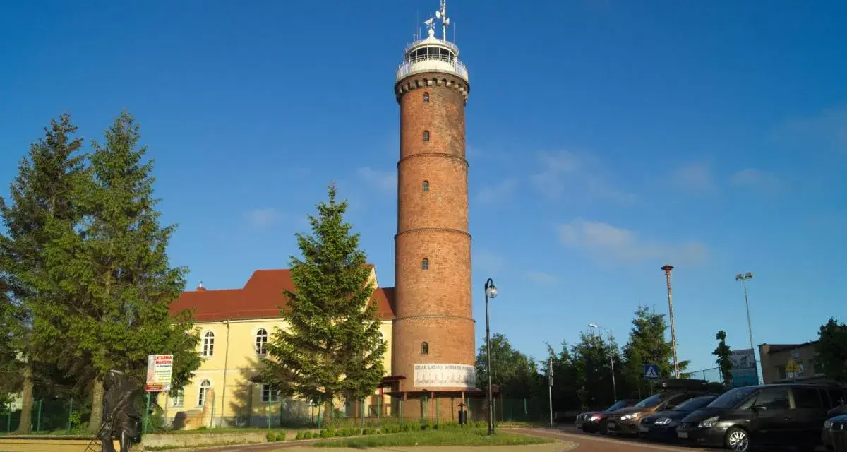 Wysoka, ceglana latarnia morska Jarosławiec góruje nad placem z drzewami i budynkiem.