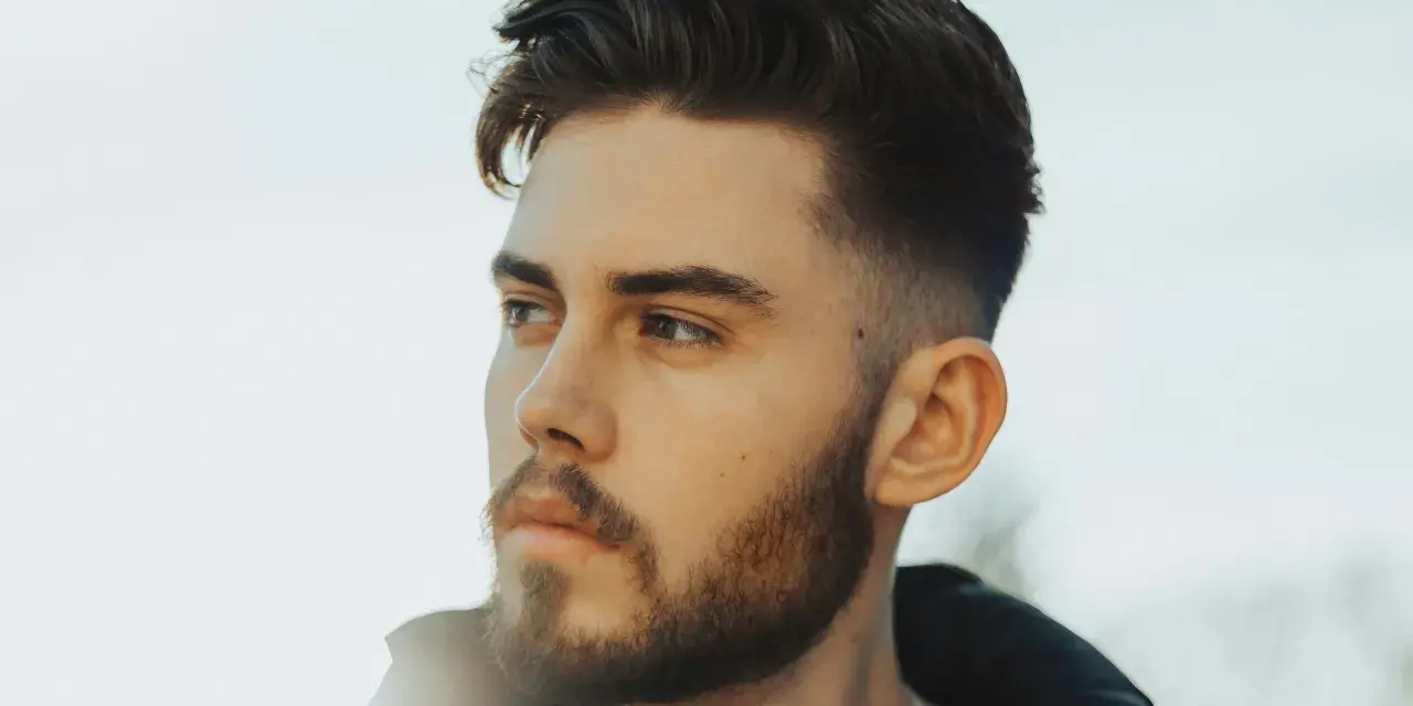 Ein Mann mit Bart und einem modernen taper fade design im Haar blickt nachdenklich zur Seite.