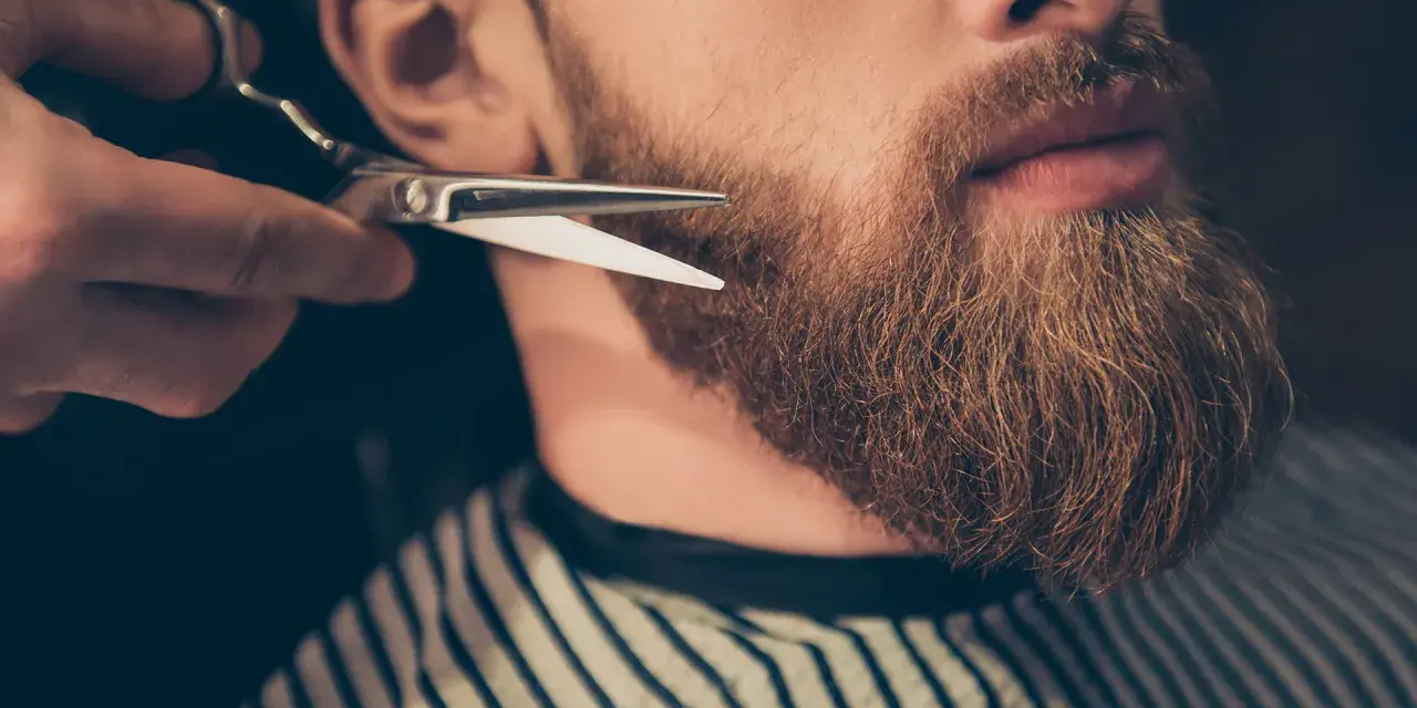 Un barbero recorta una barba larga y rojiza con tijeras.