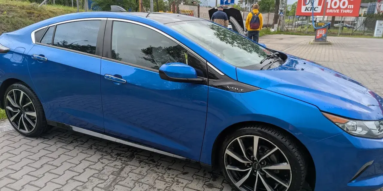Błękitny Chevrolet Volt z czarnym dachem, zaparkowany na mokrej kostce brukowej. W tle widać stację KFC.