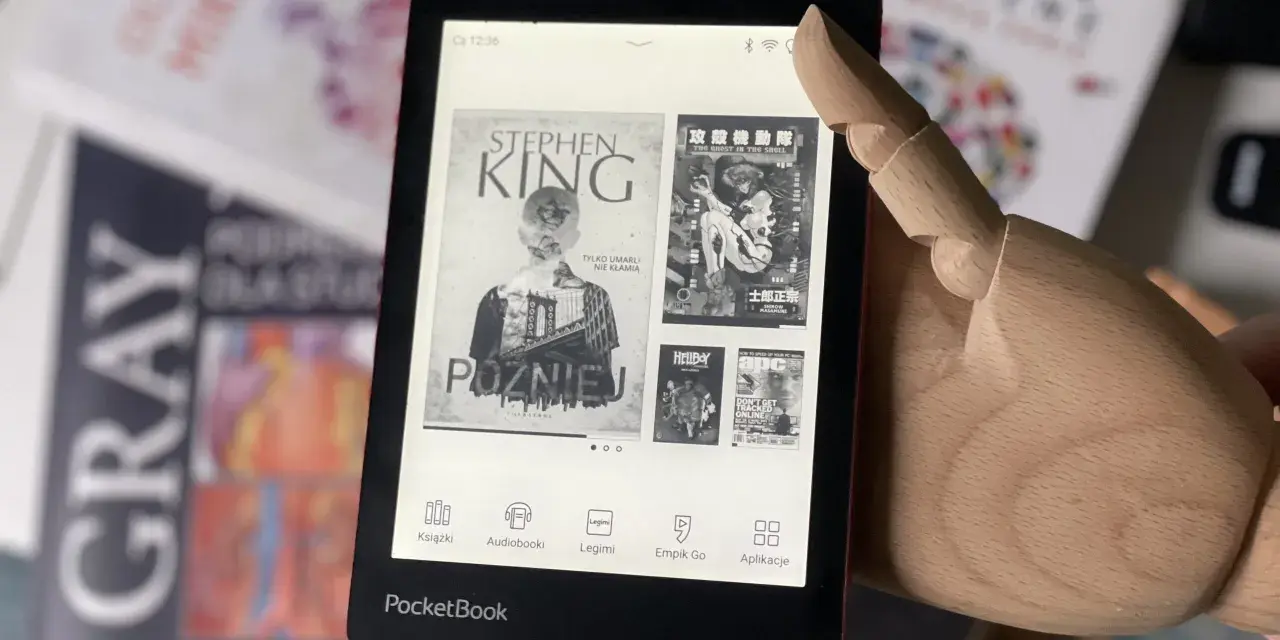 Drewniana dłoń wskazuje na ekran czytnika PocketBook, gdzie wyświetla się okładka książki Stephena Kinga "Później". W tle widać inne książki.
