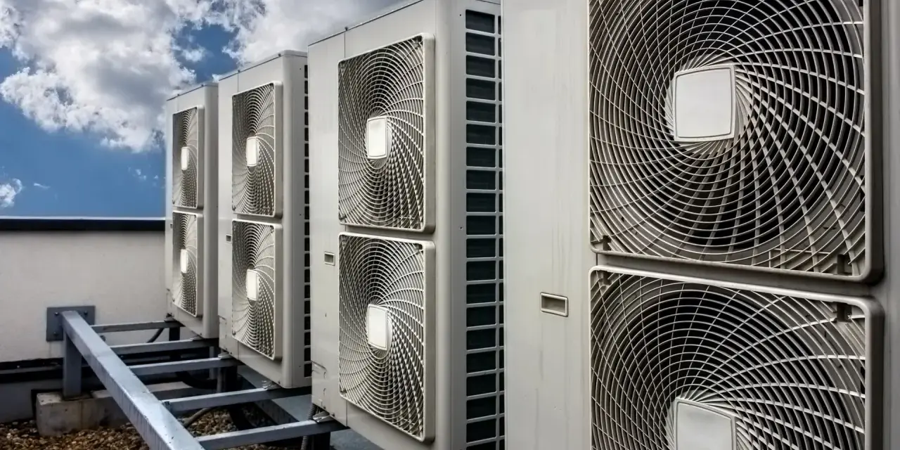 Rząd jednostek klimatyzacyjnych na dachu pod błękitnym niebem z chmurami.