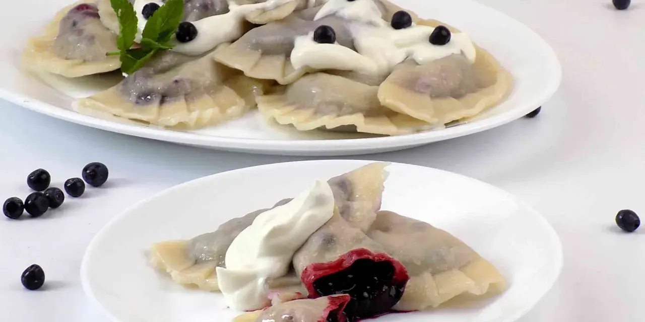 Pyszne pierogi z jagodami, polane śmietaną i ozdobione miętą. Na talerzu widać też kilka świeżych jagód.