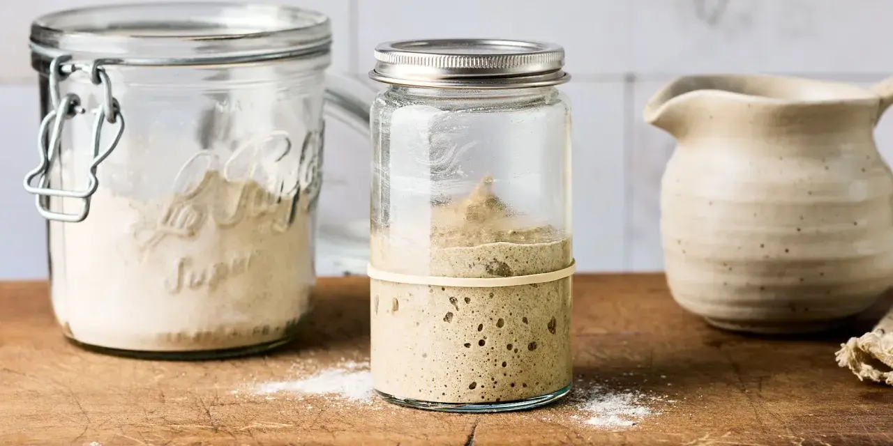 Blubbernder Sauerteig im Glas, bereit für schnelles Sauerteig herstellen. Daneben ein Glas mit Mehl und ein Krug.