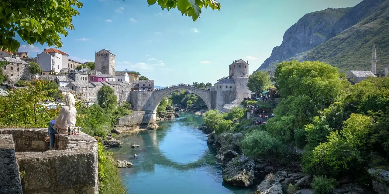 Widok na Mostar w Bośni z kamiennym mostem Stary Most nad turkusową rzeką Neretwą. W tle kamienne budynki i góry.