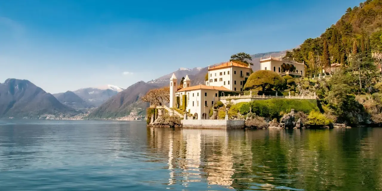 A scenic journey from Milan to Lake Como reveals this stunning villa on the water's edge, with mountains in the distance.