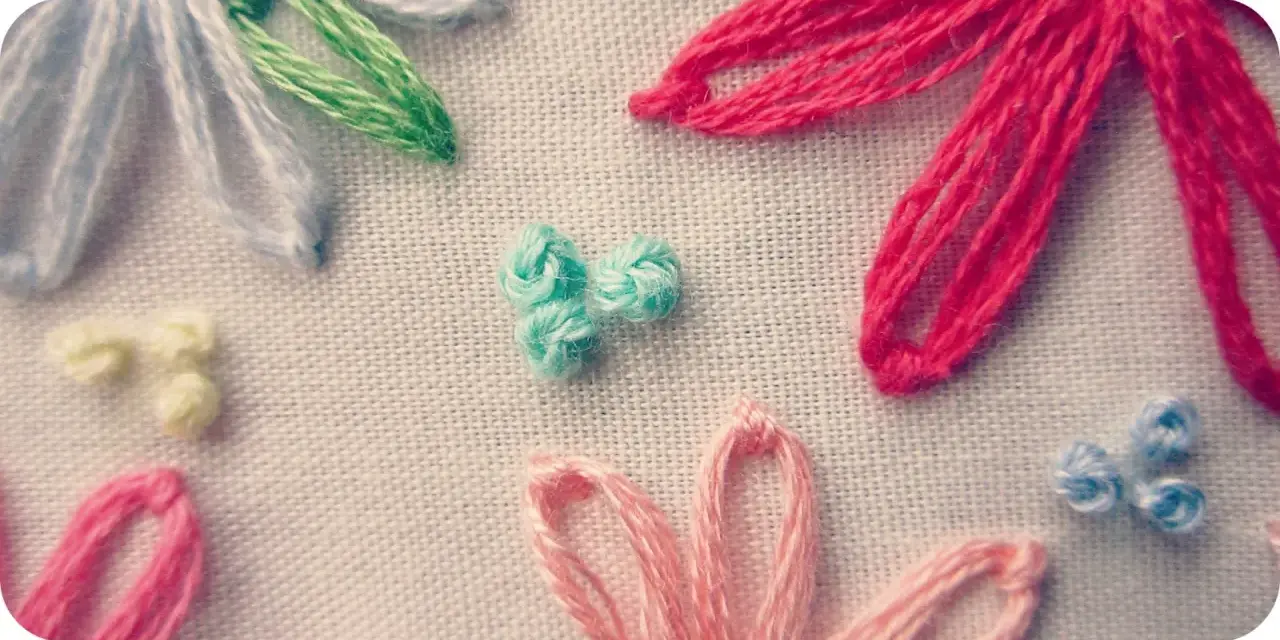 Detalle de bordado con flores de hilo. Se aprecian pétalos en rosa, rojo, azul y blanco, y un centro de nudo francés bordado en turquesa.