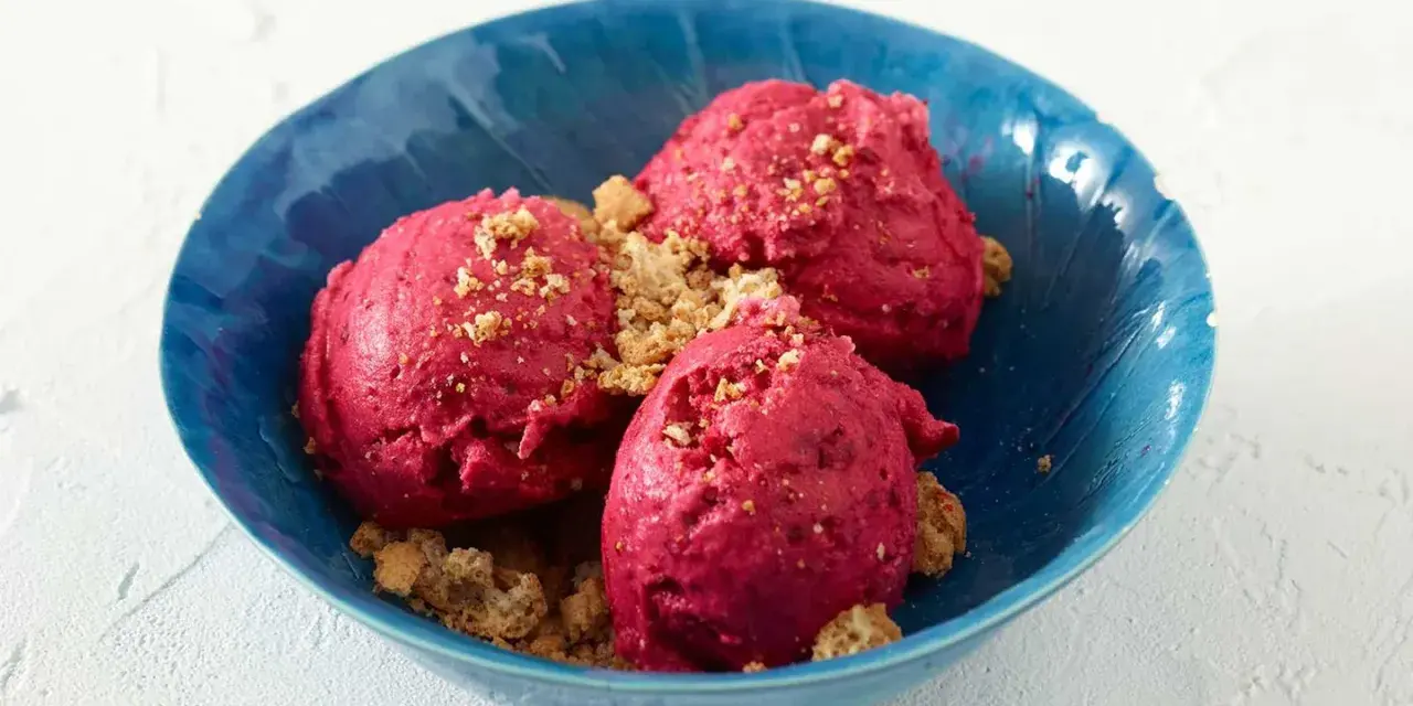 Drei Kugeln leuchtend pinkes Sauerrahm Eis, bestreut mit knusprigen Krümeln, in einer blauen Schale.