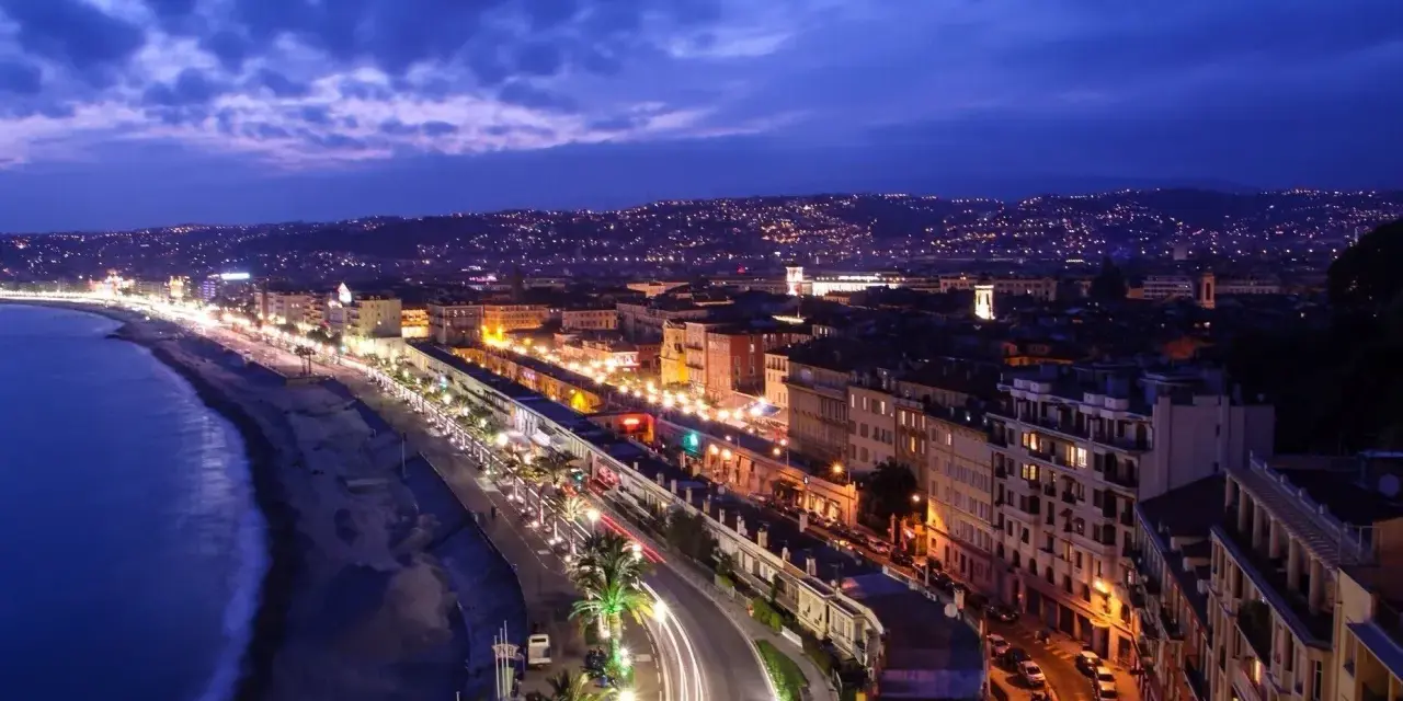 Night view of the Promenade des Anglais from Castle Hill, Nice, France, with city lights twinkling and car trails on the road.