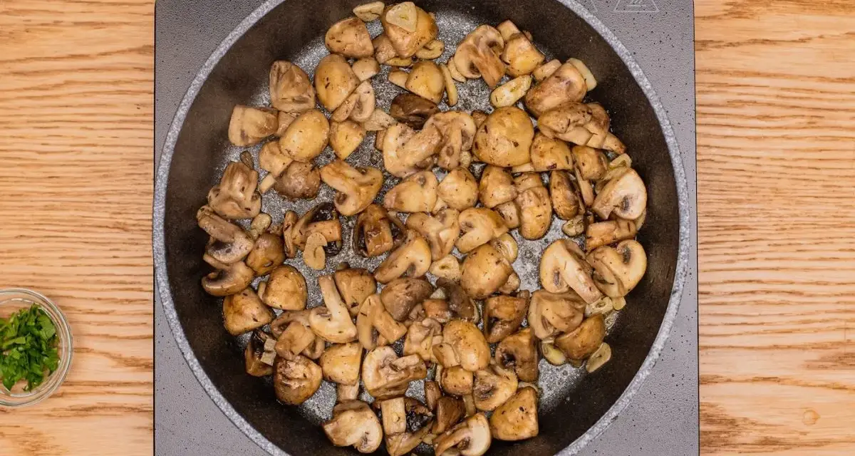 Champiñones al ajillo dorándose en sartén, con ajo y perejil picado al lado.