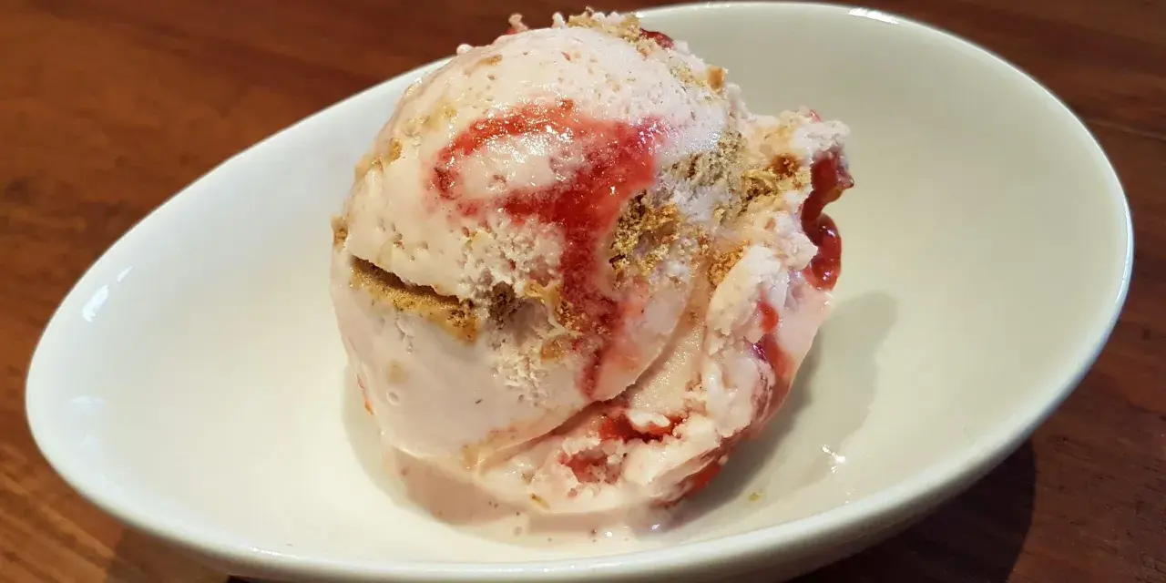 Ein Löffel cremiges strawberry cheesecake eis mit Erdbeersauce und Keksstückchen in einer weißen Schale.