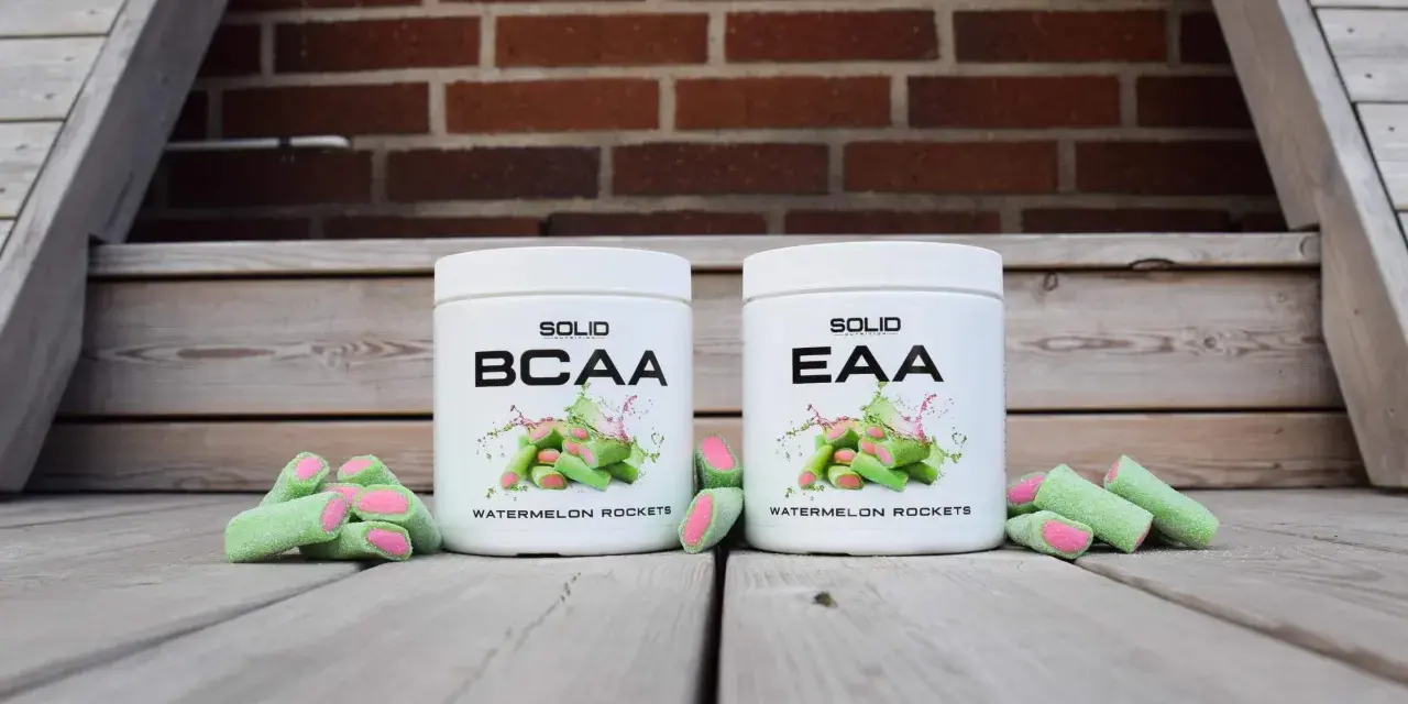 Två burkar med kosttillskott, en märkt BCAA och en EAA, står framför en tegelvägg. Runt burkarna ligger gröna godisar med rosa inuti, som ser ut som vattenmelonraketer.