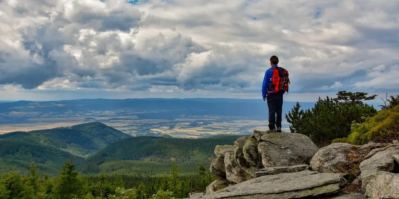 Turysta z plecakiem podziwia rozległą panoramę Gór Opawskich. Chmury na niebie tworzą dramatyczny spektakl.