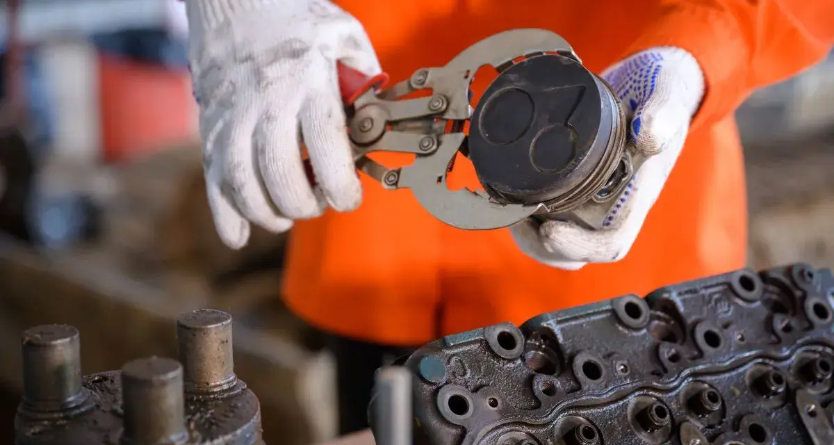 Mechanik w białych rękawicach trzyma tłok z widocznymi objawami zużytych pierścieni tłokowych. Obok leży głowica silnika.