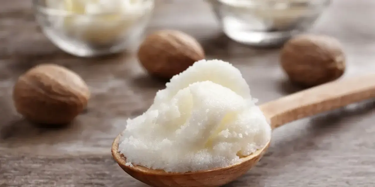 Ein Holzlöffel voller cremiger shea butter haare, daneben Nüsse und Schalen mit der Butter.
