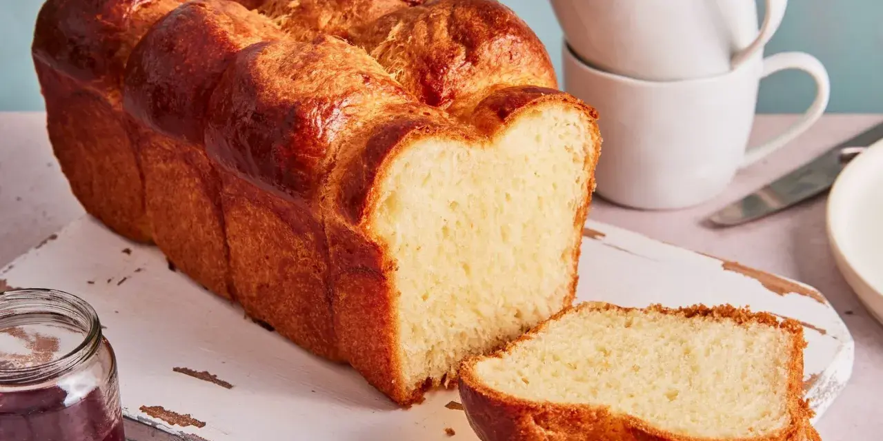 Ein goldbraunes Brioche Brot Rezept, aufgeschnitten auf einem Holzbrett mit Marmelade und Tassen im Hintergrund.