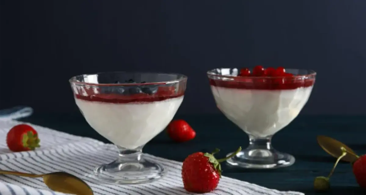 Dwie panna cotta: jedna na śmietance z borówkami, druga na jogurcie z malinami. Obok świeże truskawki i złote łyżeczki.