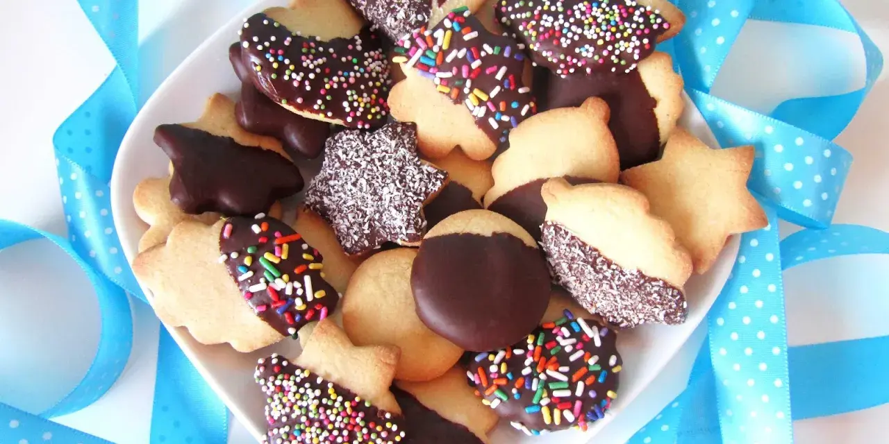 Un plato lleno de pastas de té caseras, algunas cubiertas de chocolate y chispas de colores, otras con coco rallado, rodeadas de cintas azules.