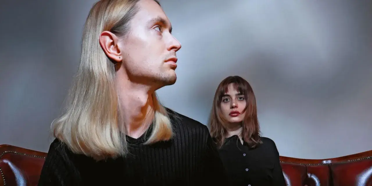 Łukasz Rozmysłowski z długimi blond włosami i kobieta z brązowymi włosami siedzą na skórzanej kanapie.