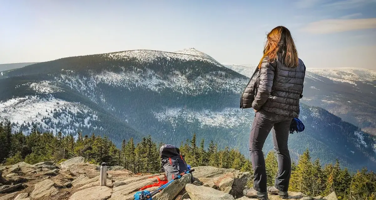 Kobieta na skalnym stole podziwia zimowy krajobraz gór. Obok niej plecak i kijki trekkingowe.