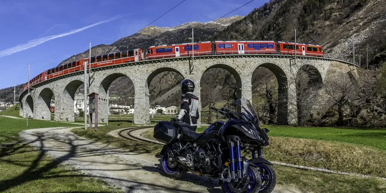 Motociclista e moto su strada sterrata, con un treno rosso che attraversa un viadotto ad archi in pietra. Paesaggio alpino con montagne sullo sfondo, vicino al passo della Novena.