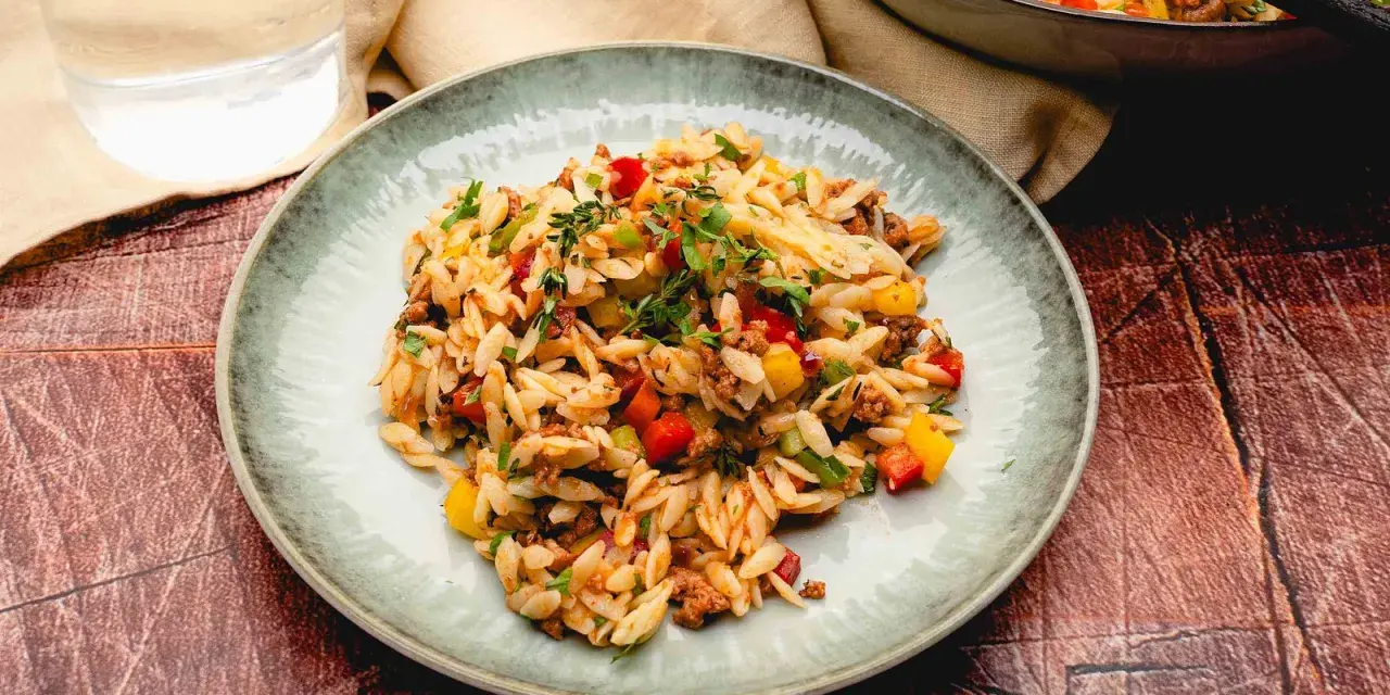 Ein Teller mit kritharaki salat mit hackfleisch, garniert mit frischen Kräutern. Daneben ein Glas Wasser und eine Pfanne mit dem Gericht.