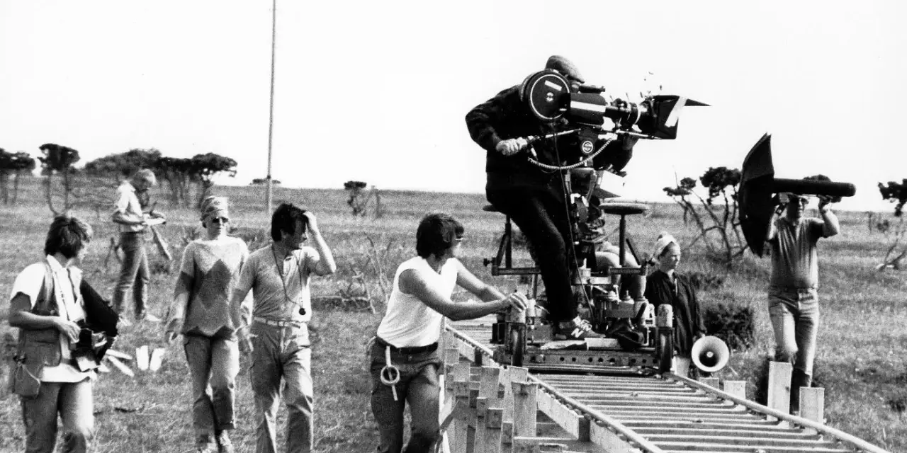 Sul set di un film, un operatore manovra una cinepresa su binari per un piano sequenza. Altri membri della troupe osservano e lavorano.