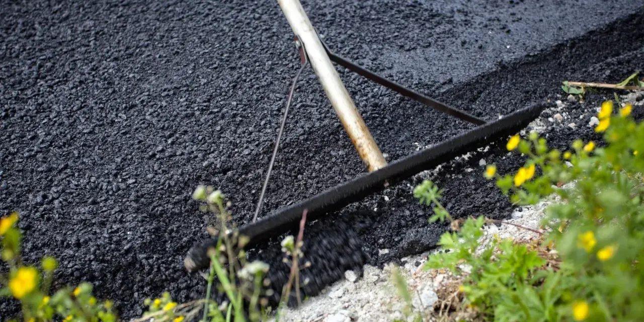 Verktyg sprider ny asfalt för att asfaltera uppfart. Grönska och gula blommor växer vid sidan.