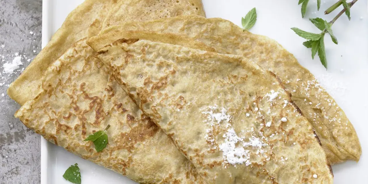 Drei goldbraune Dinkel Pfannkuchen, leicht mit Puderzucker bestäubt und mit frischer Minze garniert, auf einem weißen Teller.