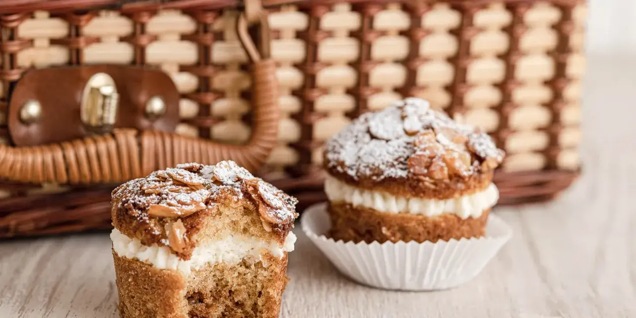 Zwei leckere bienenstich muffins mit Mandel-Topping und cremiger Füllung stehen vor einem Picknickkorb.