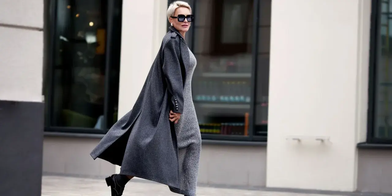 Elegante Frau ab 70 mit Kurzhaarschnitt und Sonnenbrille, die in einem langen grauen Mantel und glitzerndem Kleid durch die Stadt geht.