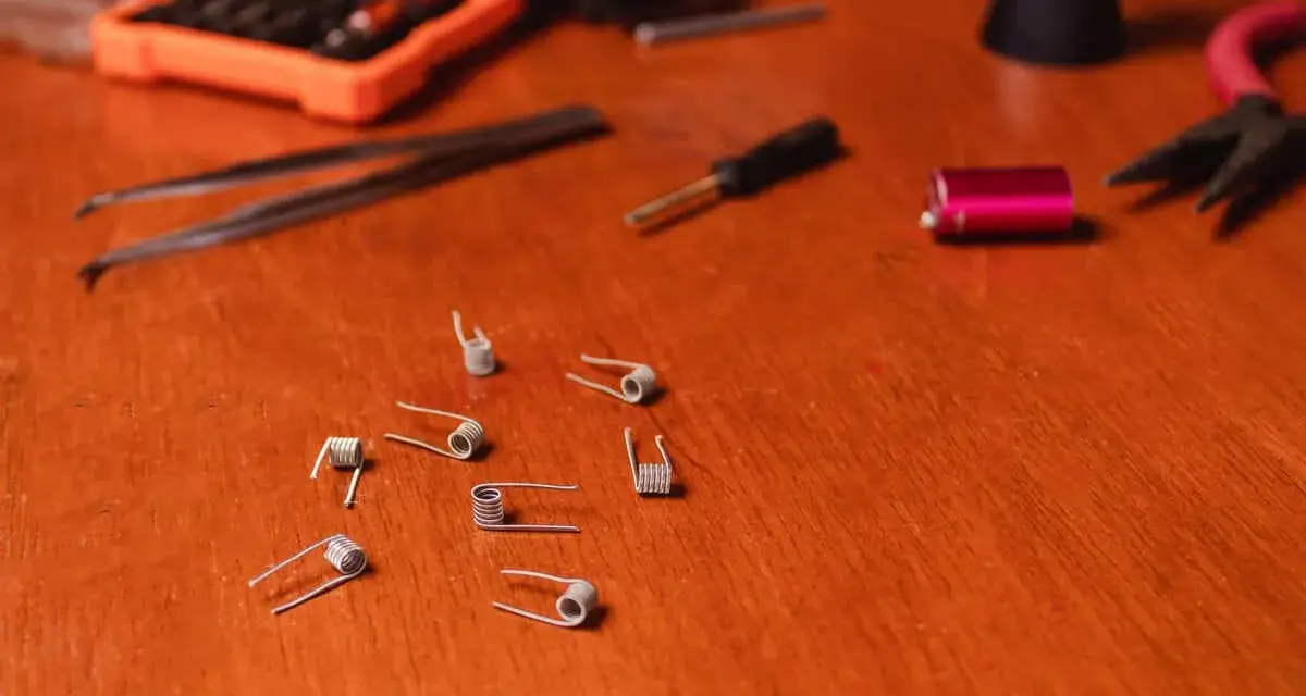 Varias resistencias comerciales de vapeo esparcidas sobre una mesa de madera, junto a herramientas como pinzas y un destornillador.