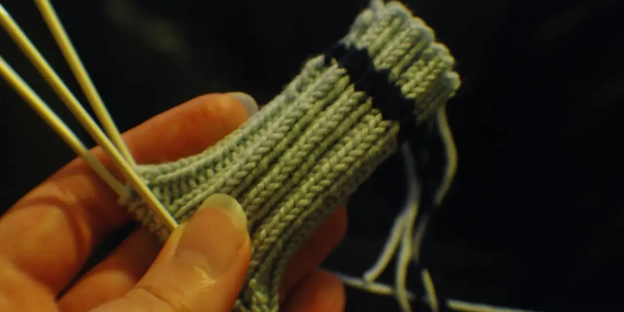En hand håller stickor och ett ljusgrönt stickat tyg med en mörk rand. Det ser ut som att någon ska sticka avig maska.