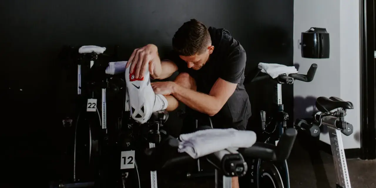 Man in black shirt stretches his outer hamstring by holding his foot, surrounded by stationary bikes in a gym.