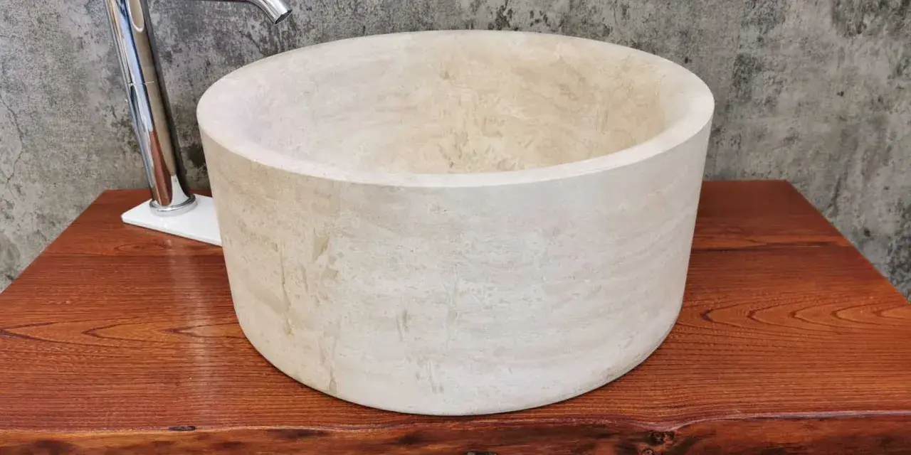 Lavabo cilindrico in pietra naturale chiara, con rubinetto cromato, su un piano in legno grezzo. Un esempio di come i nomi delle pietre naturali si trasformano in design unici.