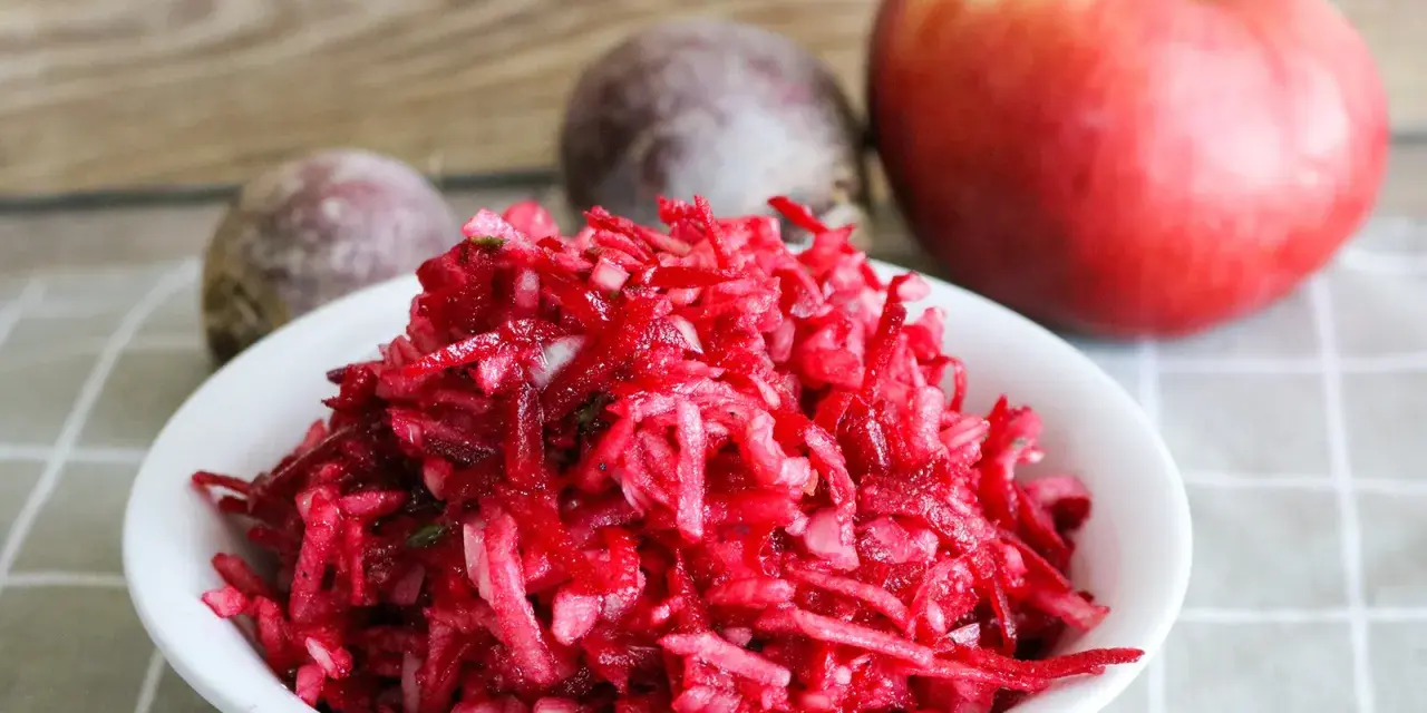 Frischer Salat aus rote Bete raspeln, mit einem Apfel und ganzen Rote Beten im Hintergrund.