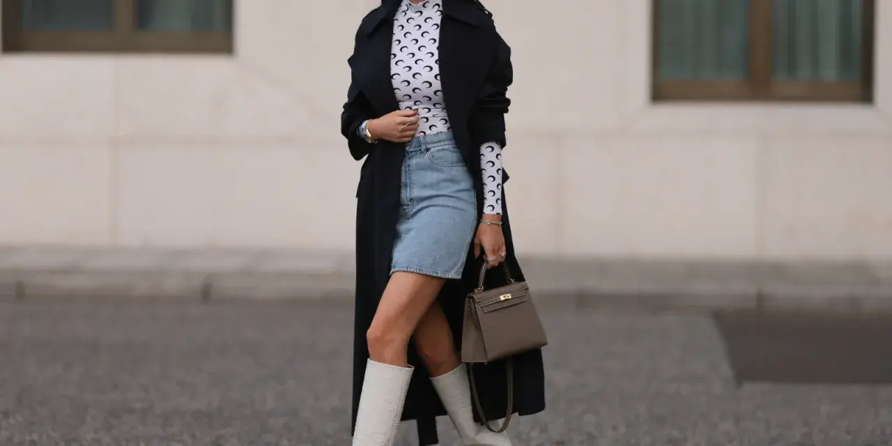 Outfit per uscire con un ragazzo: gonna di jeans, top a collo alto con stampa luna, trench nero, stivali bianchi alti e borsa grigia.