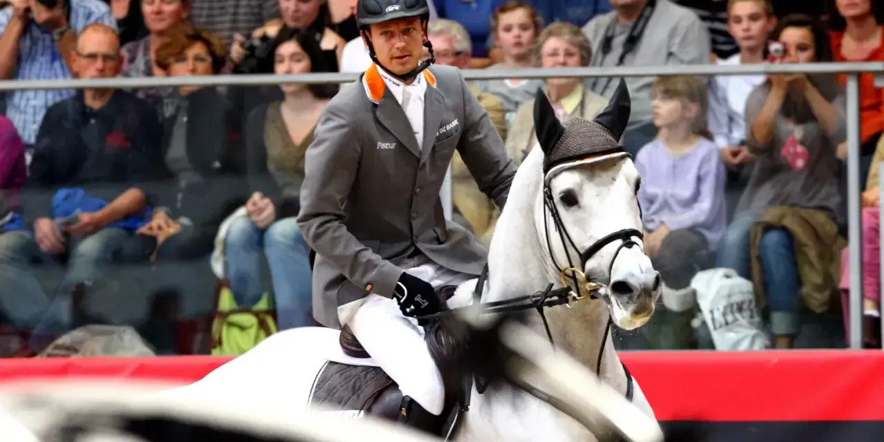 Un cavalier en tenue grise monte un cheval blanc, prêt pour la compétition. Le public est attentif.